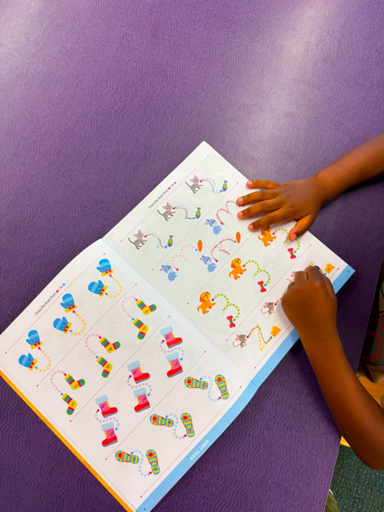 A child's hand drawing on a colorful activity book open to pages featuring various cartoon animals and patterns, with a purple textured background.