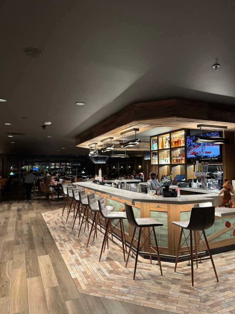 Interior view of a modern bar area with high stools, a well-lit countertop, and shelves displaying various bottles behind the bar. Fireside Restaurant at Great Wolf Lodge in Grapevine, TX. 