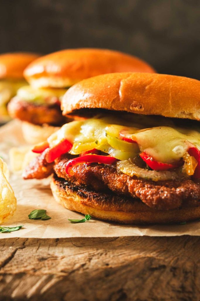 A close-up of a crispy chicken schnitzel sandwich featuring melted cheese, sautéed peppers, and onions, served on a buttery brioche bun, accompanied by a side of potato chips.