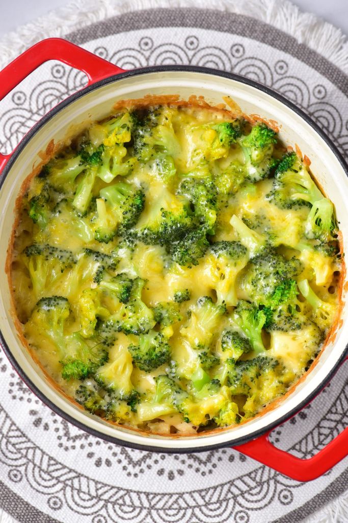 A baked broccoli and cheese casserole in a red Dutch oven, with golden melted cheese over vibrant green broccoli, placed on a decorative round placemat.