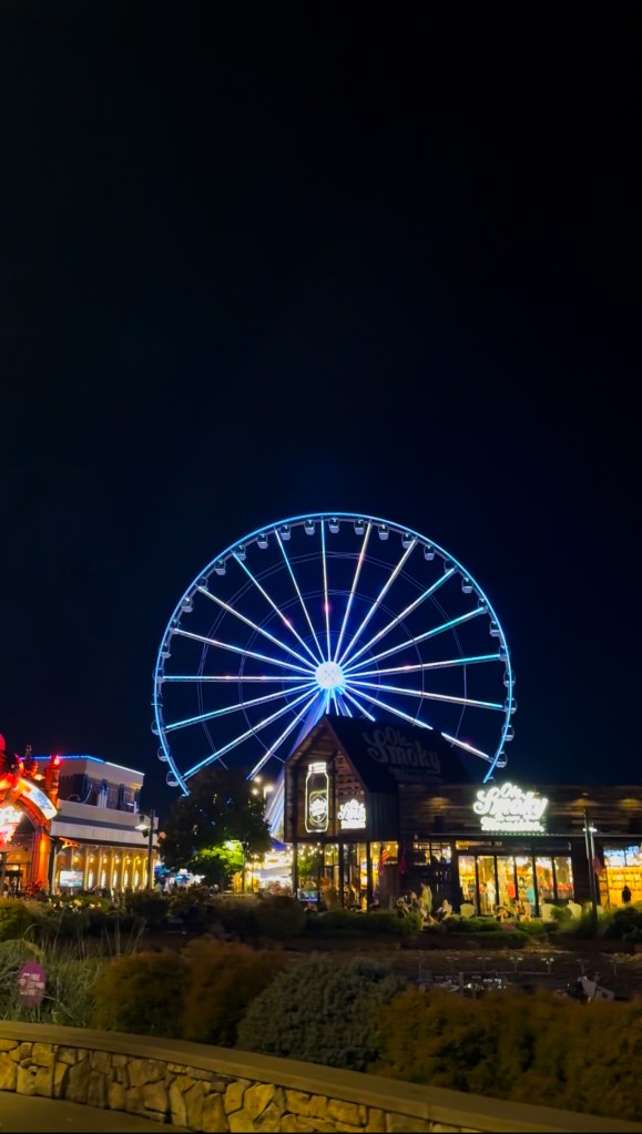 A vibrant neon-lit Ferris wheel towers over a lively street scene at night, with buildings and shops illuminated in colorful lights.