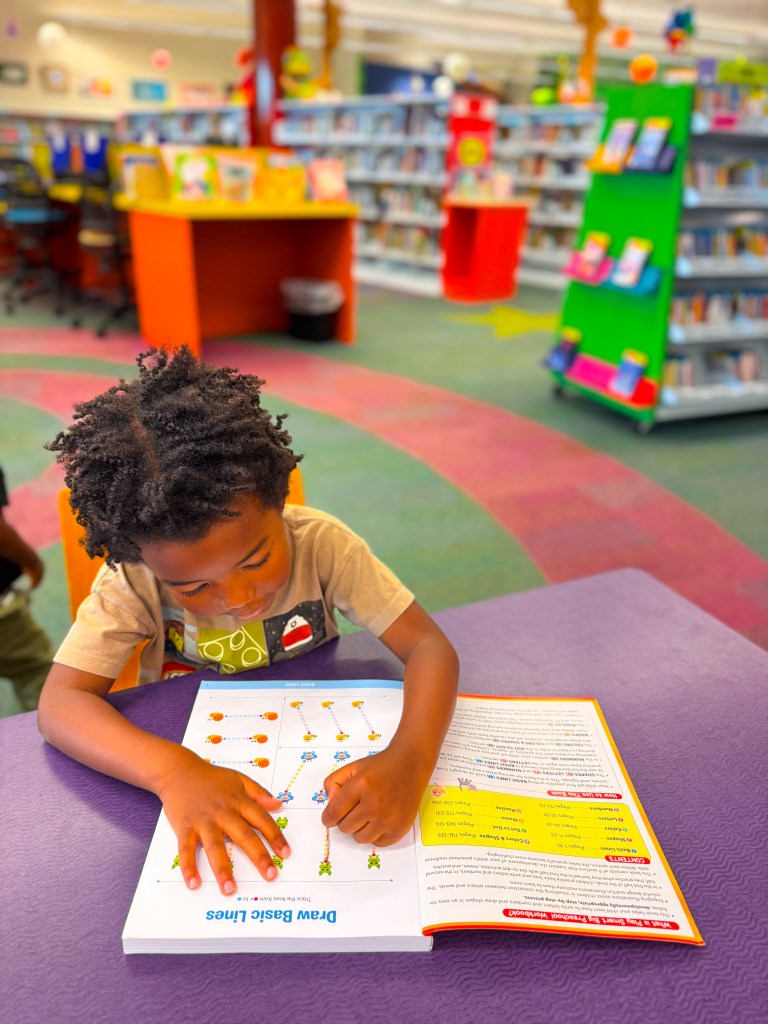 A young child with curly hair focuses on an activity book titled 'Draw Basic Lines' at a library, surrounded by colorful bookshelves.