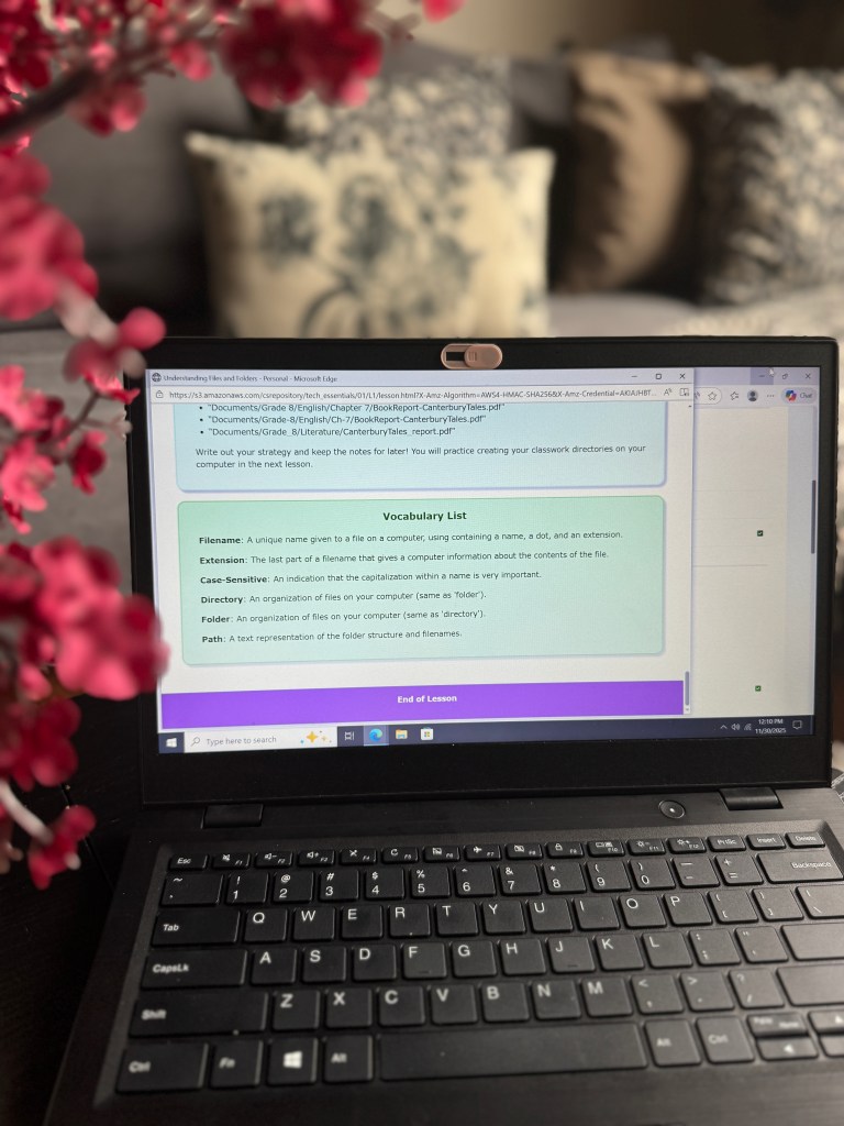 A laptop displaying an online lesson about file management, featuring a vocabulary list that includes terms like 'Filename,' 'Case-Sensitive,' and 'Directory,' surrounded by pink flowers.