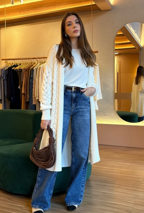 A woman stands in a clothing store wearing a cream cardigan over a white top, paired with high-waisted blue jeans. She holds a brown handbag and poses by a green sofa, with clothes displayed in the background.