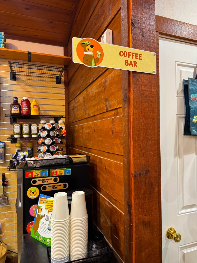 Sign for 'Coffee Bar' featuring a cartoon character holding a coffee cup, displayed in a rustic wooden interior with various coffee supplies and condiments in the background.