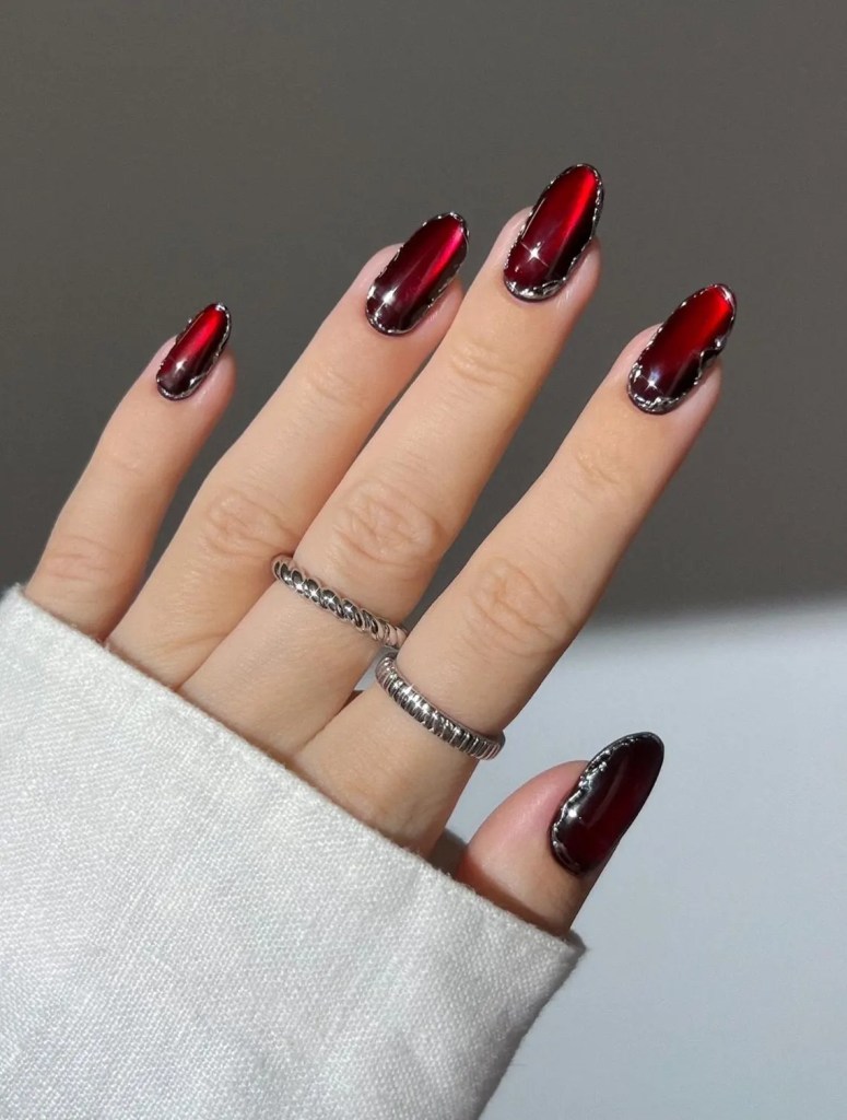 A close-up view of a hand showing manicured nails with a glossy red finish and a metallic edge, wearing two silver rings. The hand is partially covered by a white sweater sleeve.