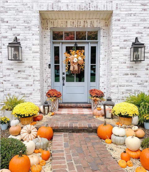 A cozy fall porch featuring a light blue door adorned with a colorful wreath. The entrance is decorated with a variety of pumpkins and vibrant mums in planters, creating a warm and inviting atmosphere.
