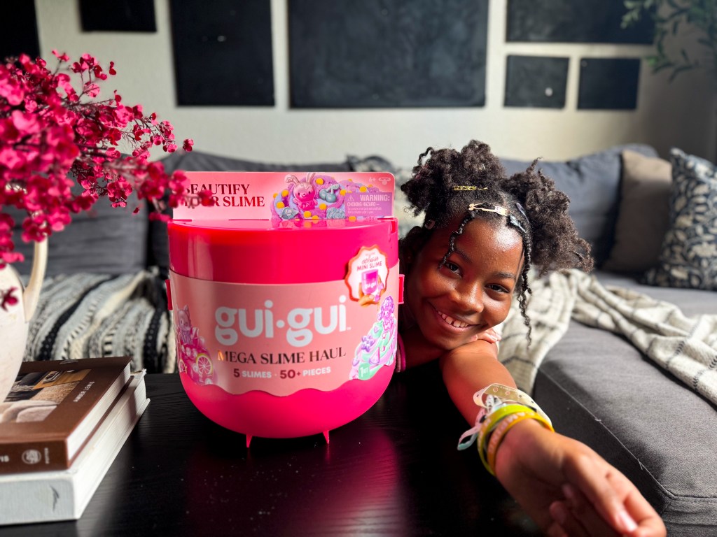 Tween girl smiling beside an open Gui Gui Mega Slime Haul case with slime creations, tools, decorations and accessories displayed on the table.