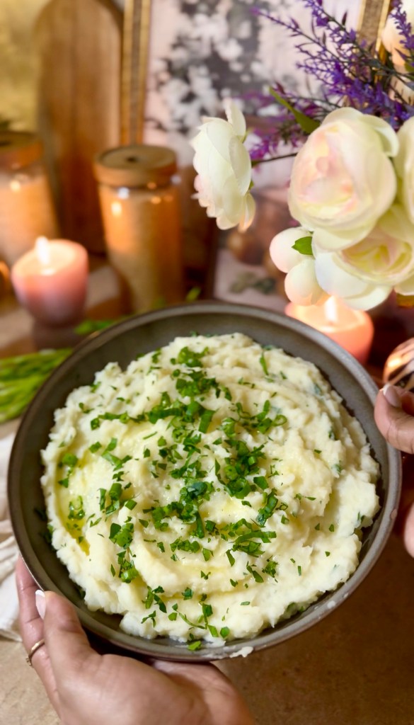 A bowl of creamy mashed potatoes topped with chopped herbs, held by hands with decorative flowers and candles in the background.