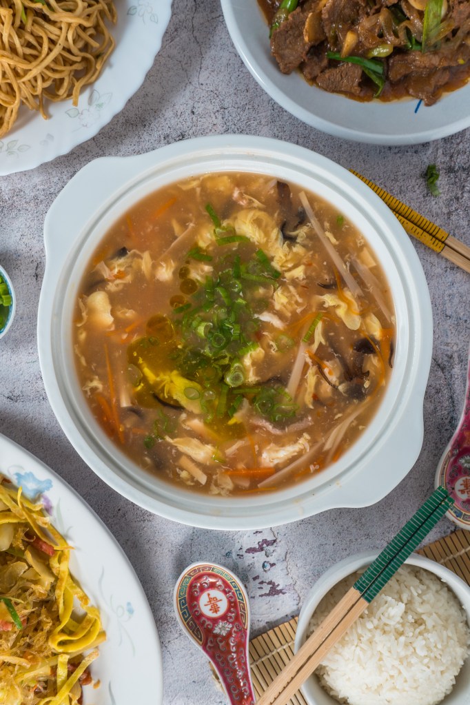 A warm bowl of soup filled with vegetables and topped with green onions, accompanied by Asian-style noodles, rice, and chopsticks, on a textured surface.