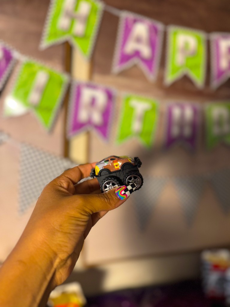 Hand holding a small monster truck toy in front of Monster Jam birthday party decorations and happy birthday banner.