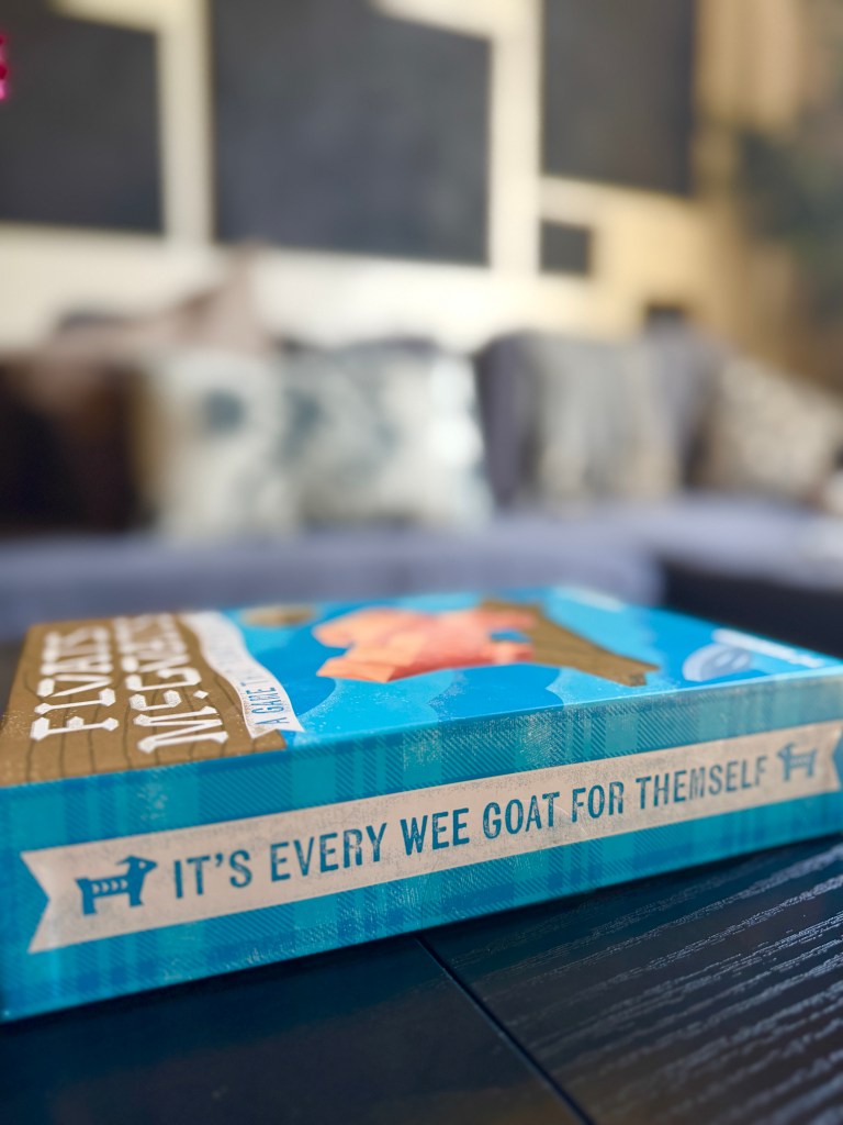 Close-up of a board game box featuring the title 'It's Every Wee Goat for Themselves' with a colorful design and a blurred background of a living room setting.