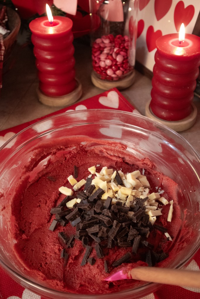 A bowl of red cake batter mixed with chopped dark and white chocolate, surrounded by festive red candles and heart decorations.