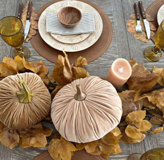 An elegant fall tablescape featuring plush pumpkins surrounded by autumn leaves, a cozy candle, and rustic dinnerware on a wooden table.