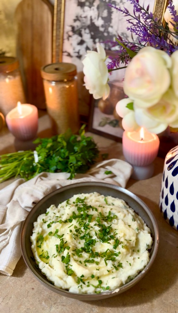 A bowl of creamy mashed potatoes garnished with chopped herbs, placed on a rustic table surrounded by candles, a bouquet of flowers, and decorative jars.