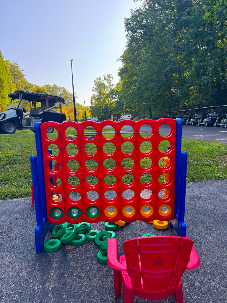A large outdoor Connect Four game featuring red and blue frames, with yellow, orange, and green game pieces scattered around. A small red plastic chair is visible in the foreground.