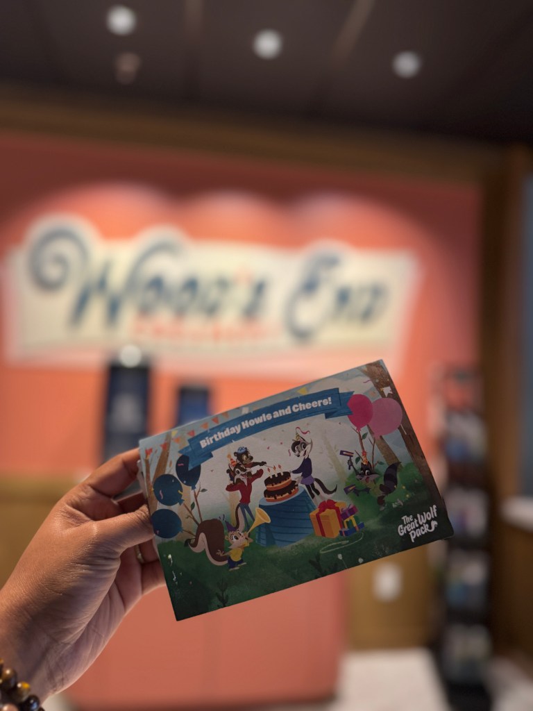 A hand holding a birthday card with a colorful illustration featuring characters celebrating a birthday, balloons, and a cake, with the Great Wolf Lodge in Grapevine, TX logo in the corner.