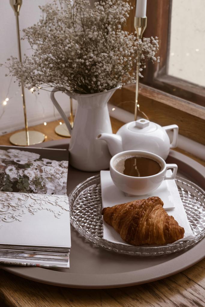 A cozy scene featuring a white teapot, a cup of coffee, and a croissant on a glass plate, accompanied by a bouquet of dried flowers in a pitcher, all arranged on a tray near a window.