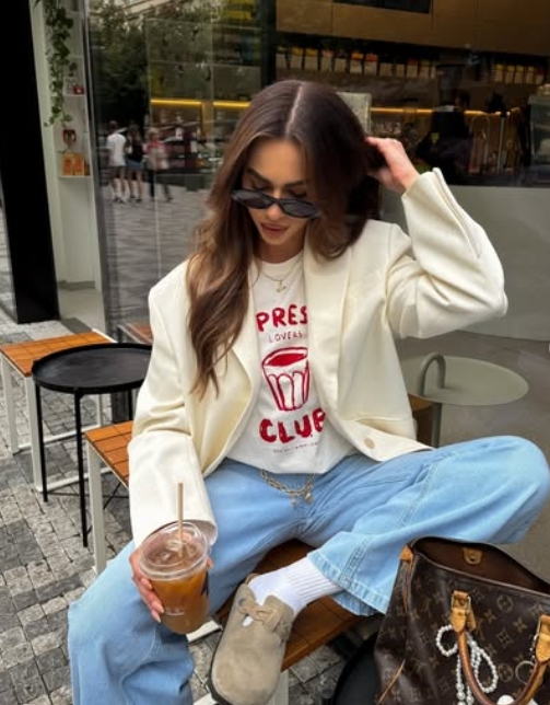 A woman sitting outdoors in a trendy outfit featuring a white blazer, graphic tee, and light blue jeans, holding a glass of iced coffee.