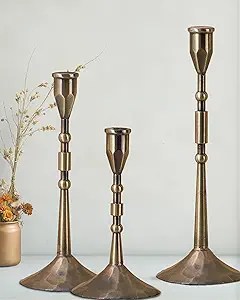 Three metallic candle holders of varying heights with a minimalist design, set against a simple backdrop decorated with dried flowers in a vase.