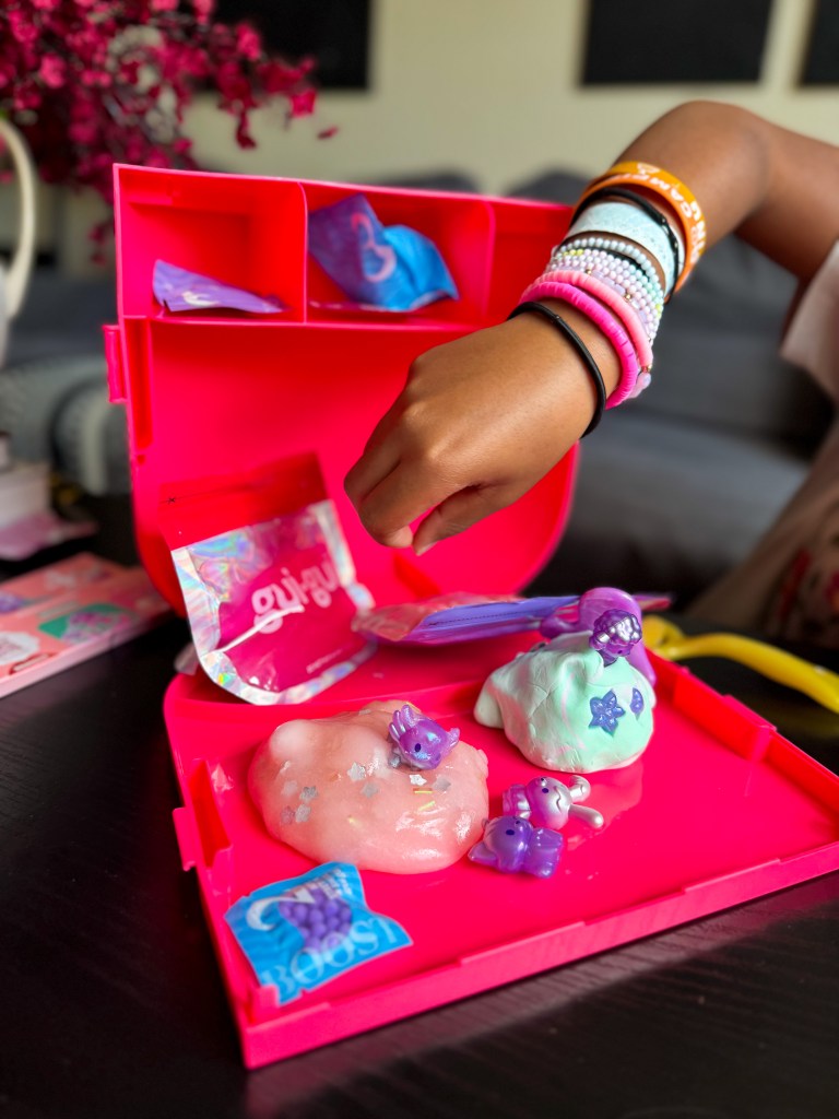 A child's hand interacting with colorful slime inside a bright pink activity kit, featuring various small molded figures and accessories.