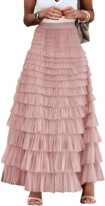 A woman wearing a tiered maxi dress in soft pink, showcasing multiple ruffles. She is accessorized with bracelets and a bag, exuding a feminine and stylish look.