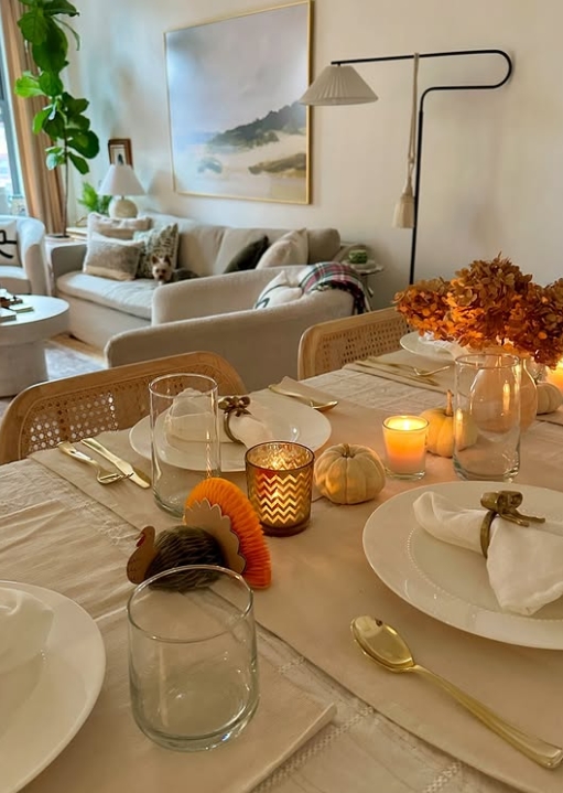 A beautifully set Thanksgiving table featuring elegant dinnerware, layered napkins, candles, and autumn-themed decorations, including small pumpkins and a decorative turkey, set in a cozy dining space.