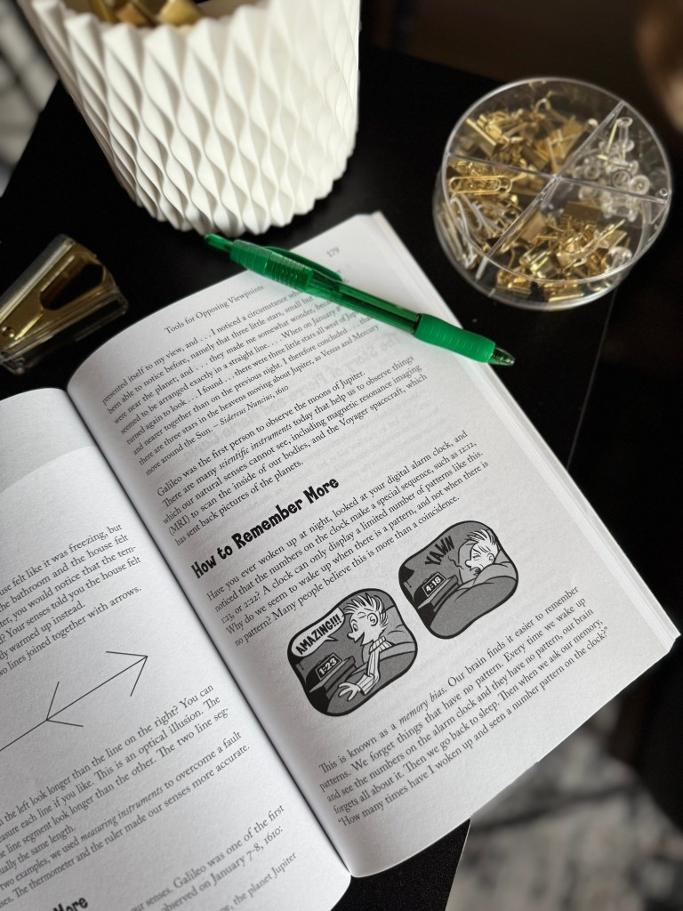 An open page from a book with illustrations and text about memory techniques, accompanied by a green pen, a decorative container, and a bowl of paper clips.