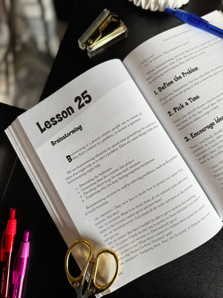 A close-up of an open book displaying Lesson 25 titled 'Brainstorming', featuring headings and instructions. Nearby, colorful pens and a pair of scissors are placed on a dark surface.
