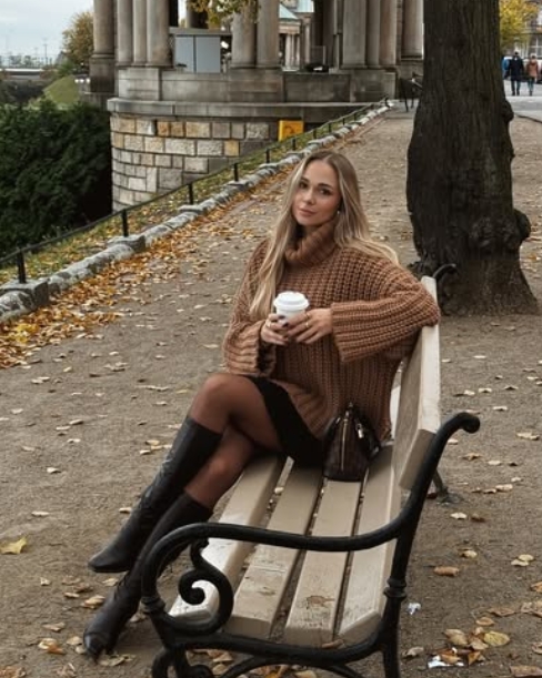 A woman sitting on a bench in a park during fall, wearing a chunky brown sweater, a black skirt, and knee-high boots. She is holding a coffee cup and has long hair down.