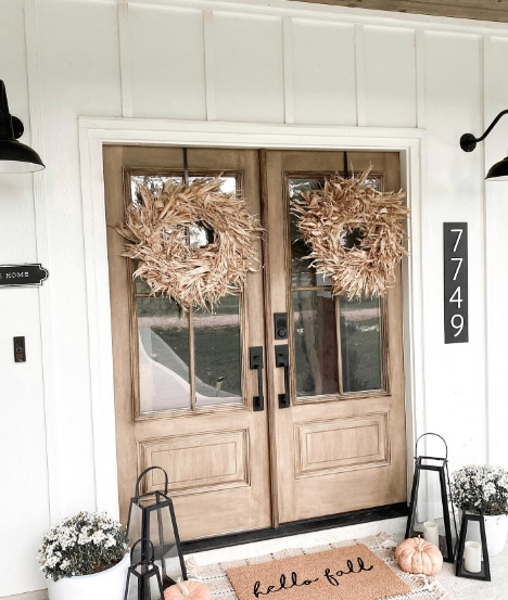 A cozy front porch featuring double wooden doors adorned with natural wreaths and a welcoming 'hello fall' doormat, complemented by lanterns and decorative plants.