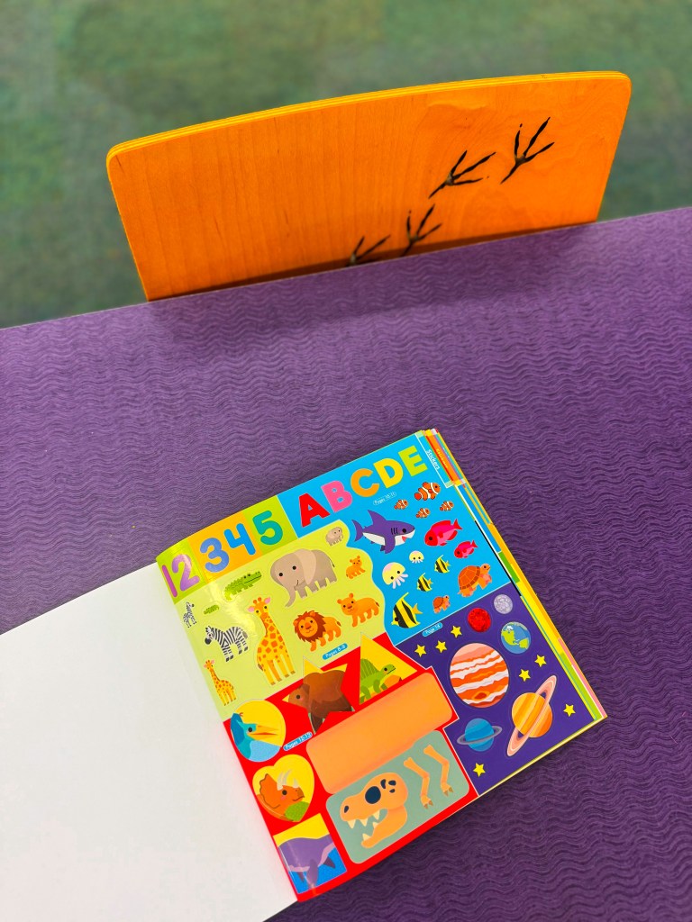 A colorful children's sticker book opened to a page with numbers and letters, featuring various animals and space-themed stickers, placed on a purple tablecloth with a wooden chair in the background.