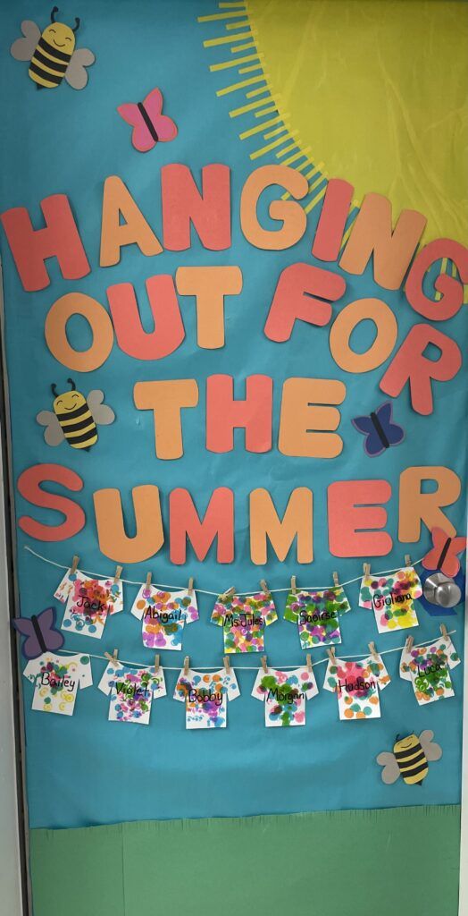 Colorful bulletin board decoration with the text 'HANGING OUT FOR THE SUMMER' featuring bees and butterflies, along with hanging art pieces labeled with names.