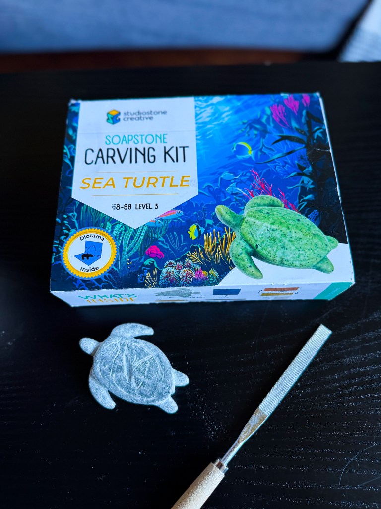 A soapstone carving kit labeled 'Sea Turtle' with a completed green turtle sculpture beside a carving tool, set against a colorful underwater background on the box.