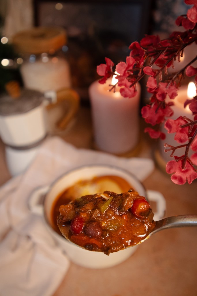 A close-up of a spoonful of hearty chili held above a white bowl of chili, with decorative flowers and candles in the background.