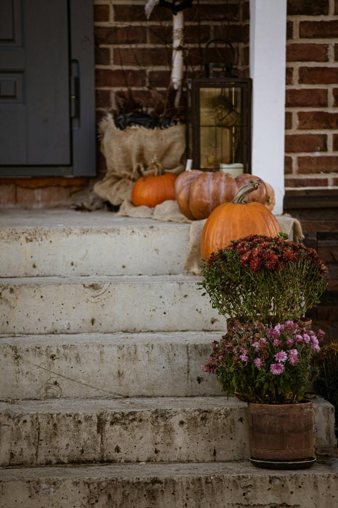 A cozy autumn scene featuring several pumpkins and potted plants on a set of concrete stairs leading up to a home entrance, with a lantern and burlap bag nearby.