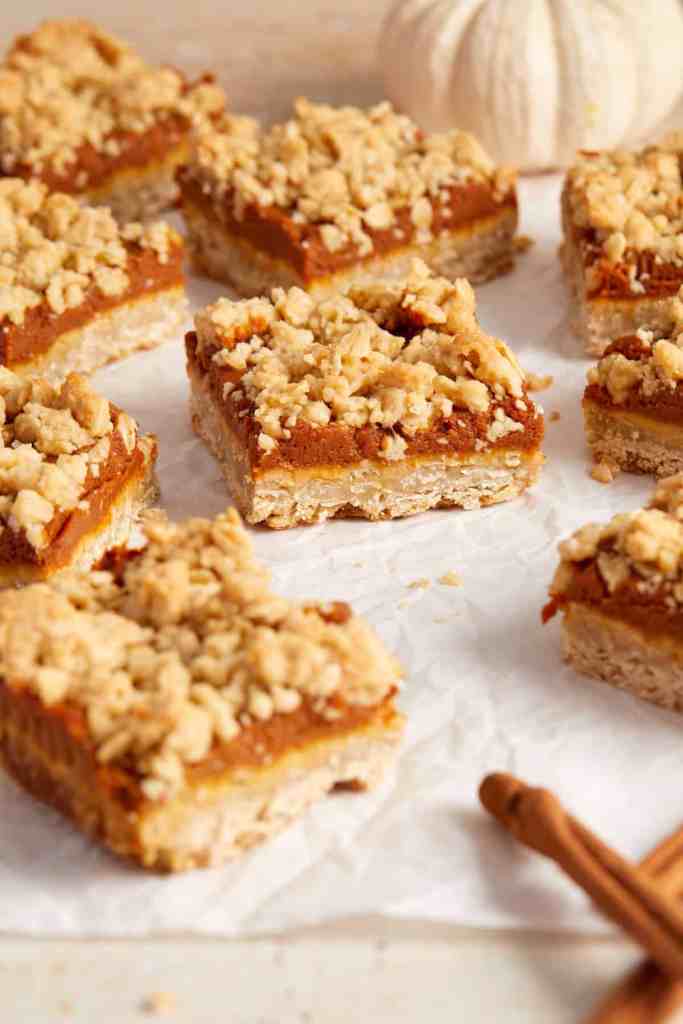 A close-up of pumpkin bars topped with a crunchy oat crumble, arranged on parchment paper, with a small white pumpkin and cinnamon sticks in the background.