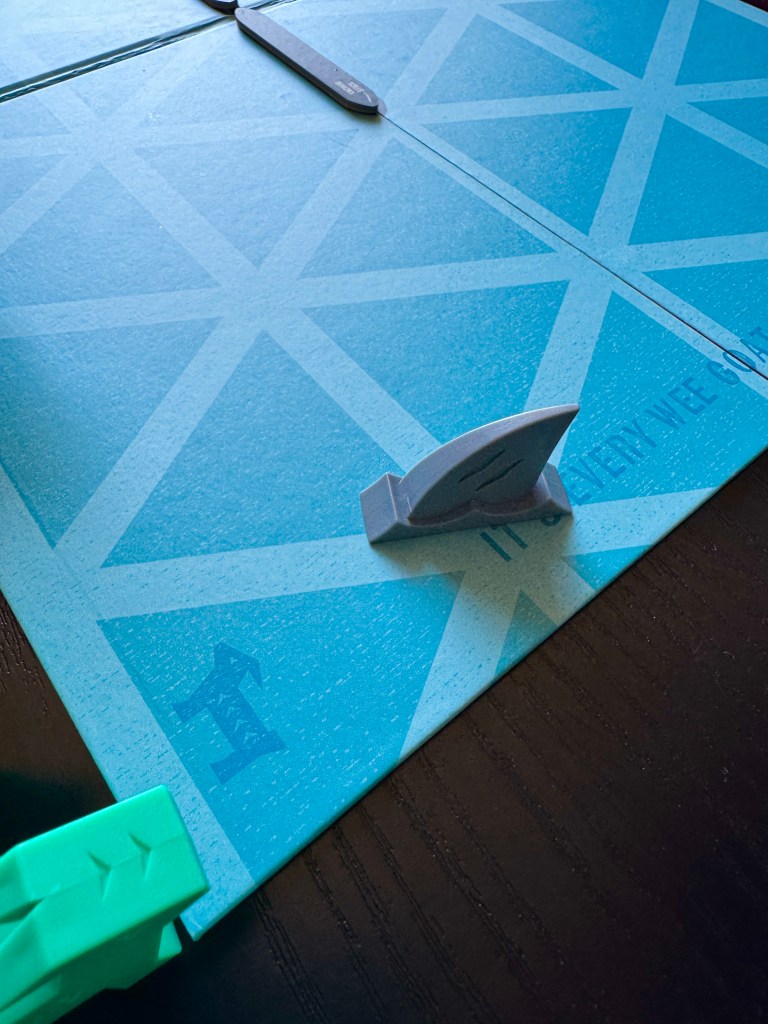 A close-up view of a game board featuring a blue surface with a geometric pattern and game pieces including a gray shark and a green piece.
