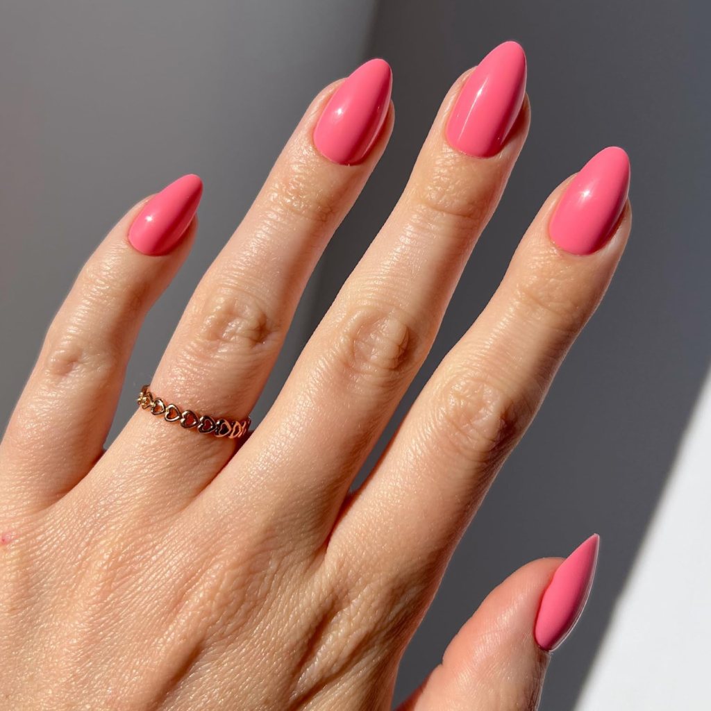 A close-up of a hand with manicured, glossy pink almond-shaped nails and a delicate gold ring on the ring finger.