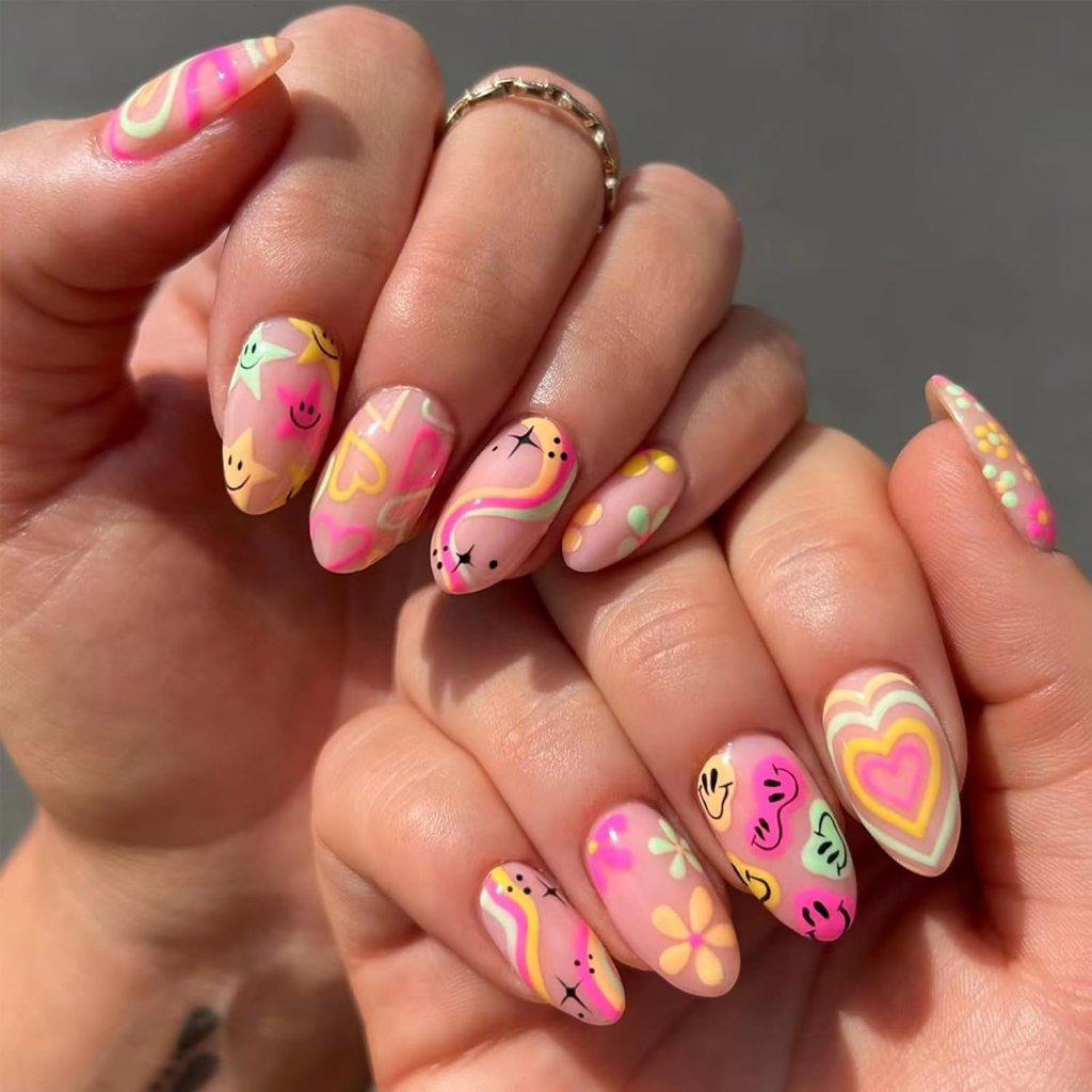 Close-up of hands showing colorful, patterned nails with designs including hearts, flowers, and smiley faces on a nude background.