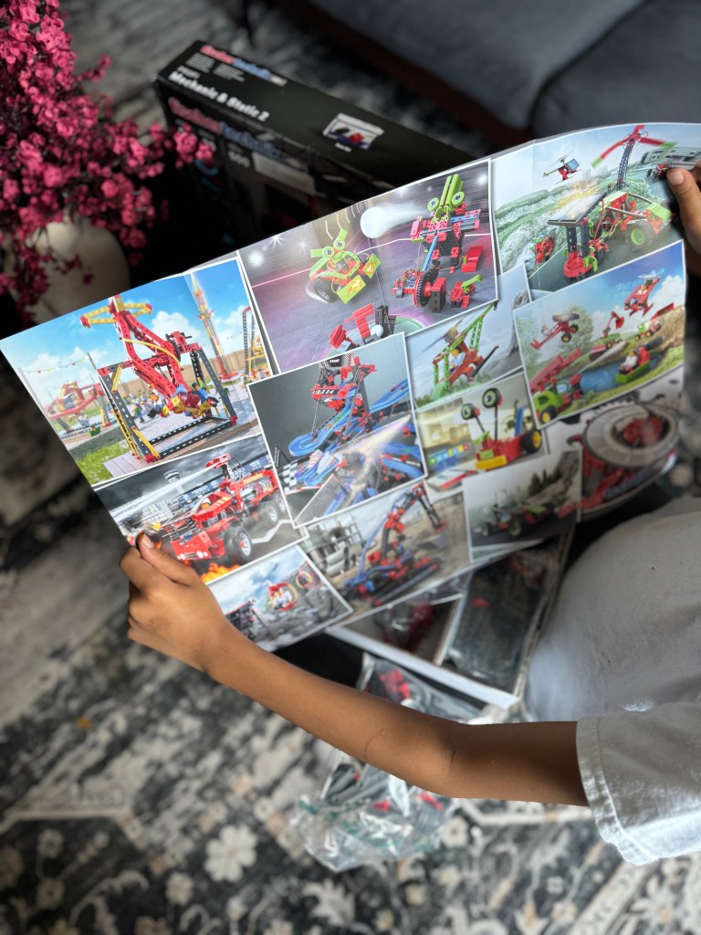 A child holding an instruction booklet while unpacking the Fischer Technik STEM Kit, with building components visible in the background.