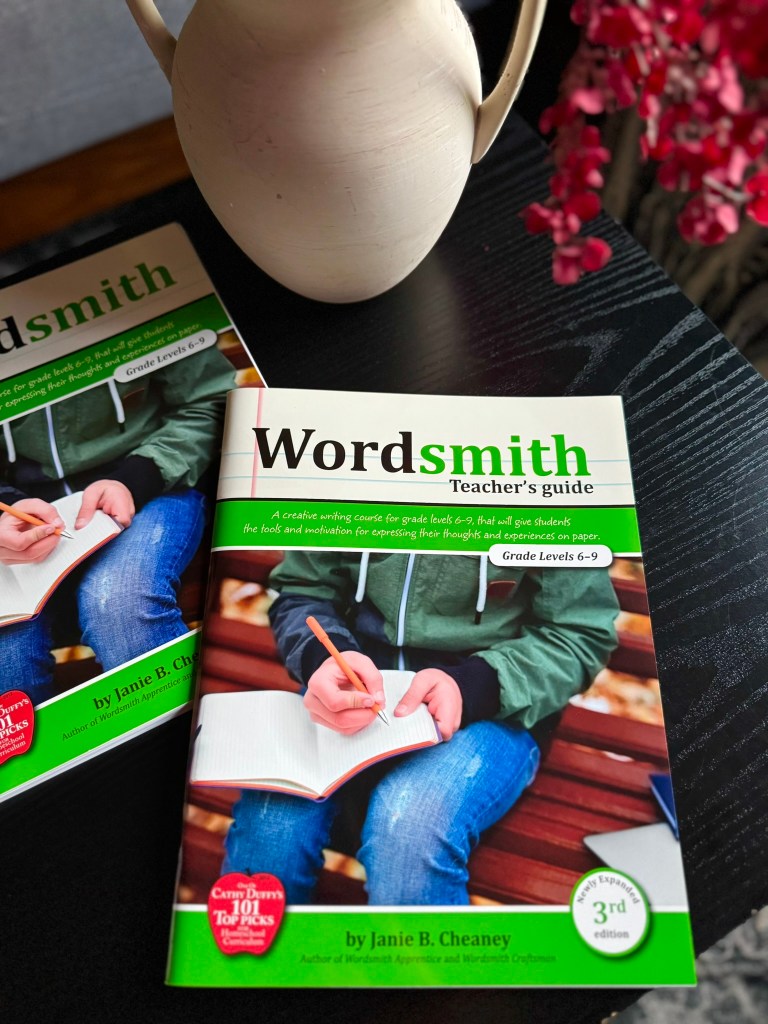 A close-up of a teacher's guide book titled 'Wordsmith' resting on a table, with a person taking notes in a notebook. A cream-colored pitcher and a brown decorative background are also visible.