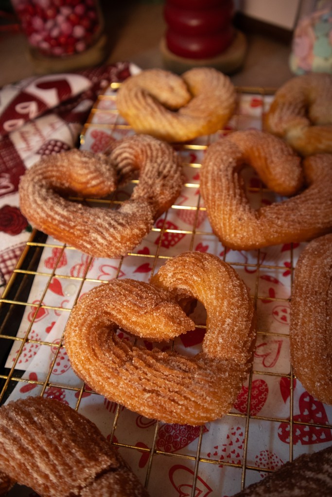 A tray of heart-shaped churros dusted with sugar, arranged on a cooling rack. The background features a patterned cloth and jars filled with colorful candies.