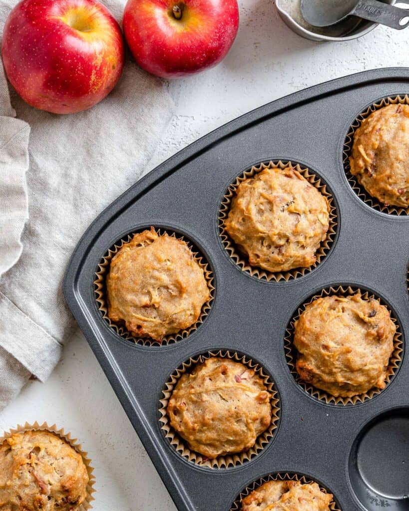 Baked muffins in a tray, surrounded by two red apples and a measuring cup.