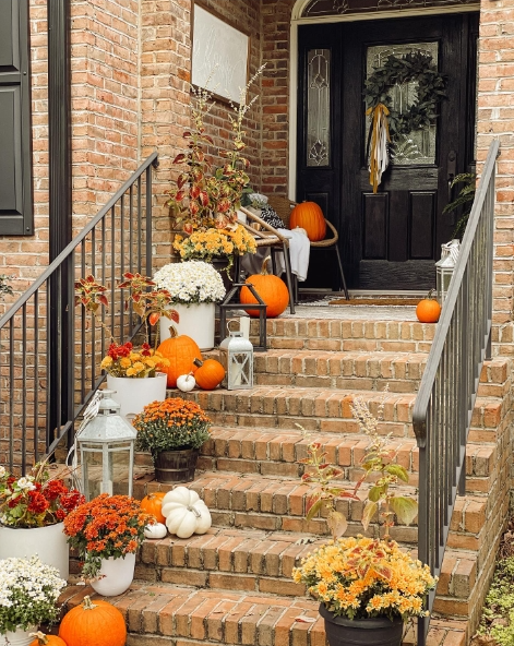 A cozy fall porch scene featuring brick steps adorned with various pumpkins, vibrant flowers, and decorative lanterns. The inviting black front door is complemented by seasonal foliage, creating a warm autumn atmosphere.