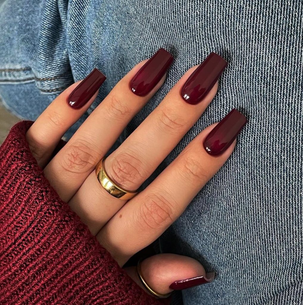 A close-up of a hand showing well-manicured nails painted in a deep burgundy color, wearing a gold ring, with a cozy burgundy sweater sleeve visible and denim fabric in the background.