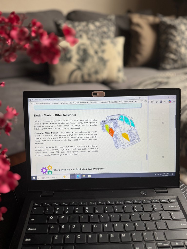 A laptop displaying a lesson on Computer Aided Design (CAD), featuring text about design tools in various industries, with a floral arrangement in the foreground and a cozy living room background.