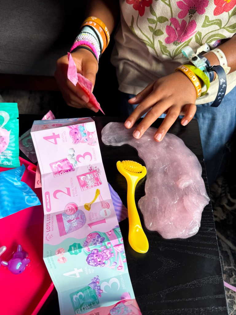 A child's hands are seen working with colorful materials on a dark table, including a pink slime-like substance, a yellow tool, and brightly colored packaging featuring instructions.