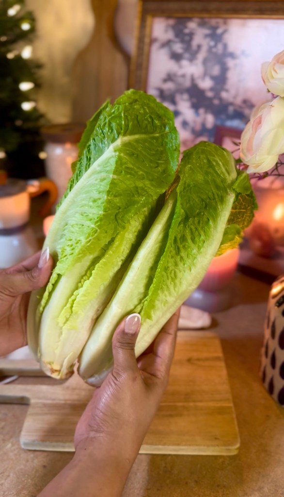 A person holding two heads of romaine lettuce in their hands, with a wooden cutting board and candles in the background.
