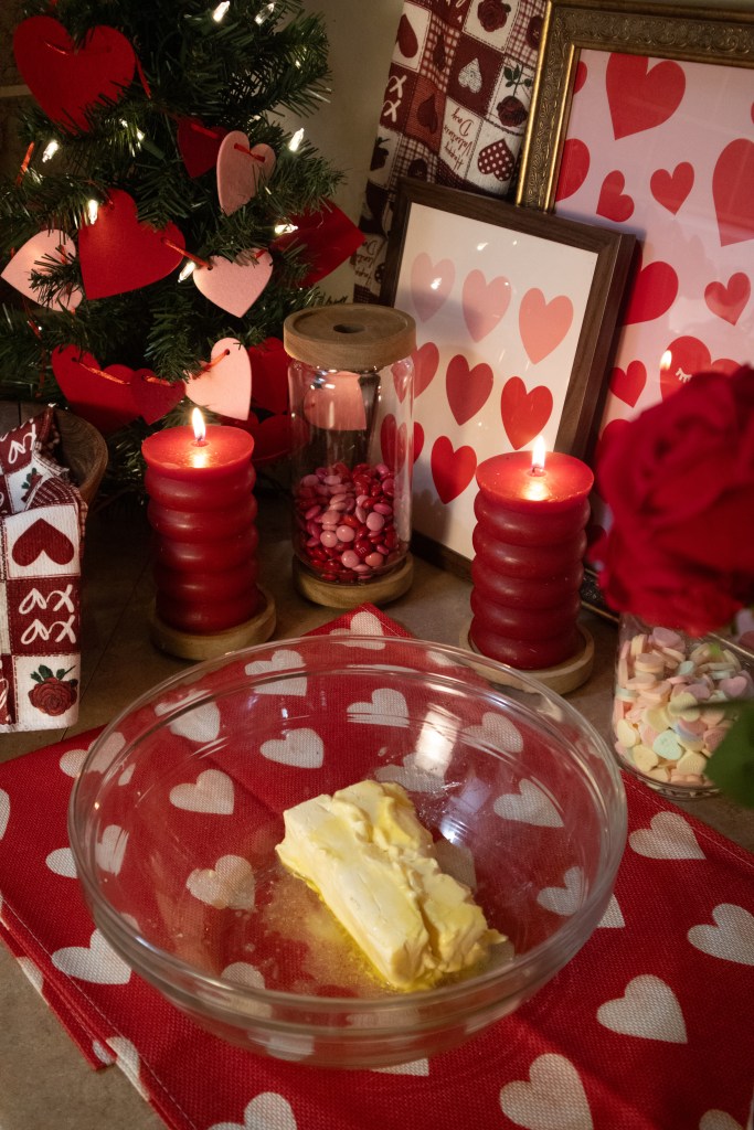 A festive setup featuring a small Christmas tree adorned with heart decorations, two red candles, and a bowl of butter on a heart-patterned tablecloth. Other decorative elements include heart-themed artwork and candy in glass containers.